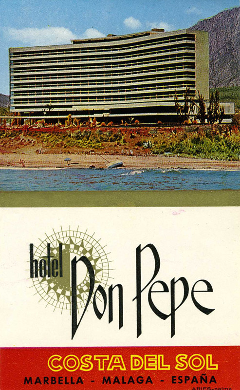 View of waterfront hotel with beach surrounded by mountains; red, blue, gold
