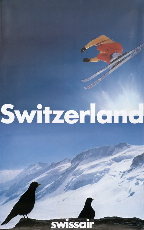 Skier soaring in sky, mountains behind, two birds in foreground; blue, white, red, black
